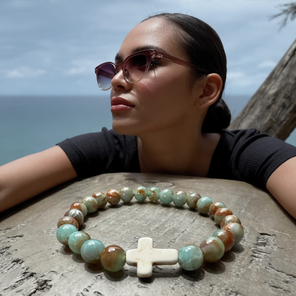 Salt and Light Merch Christian bracelet on young Hispanic woman by the ocean – mint green and brown ombre gemstone beads with a white ceramic cross centerpiece. Stylish faith jewelry lifestyle photo.
